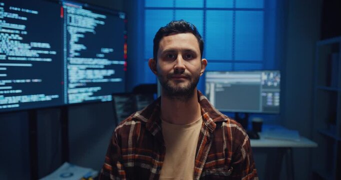Portrait of confident Caucasian male programmer smiles looking at camera in office at night. Professional developer workspace, Secure computer network system, Advanced cybersecurity data concept.