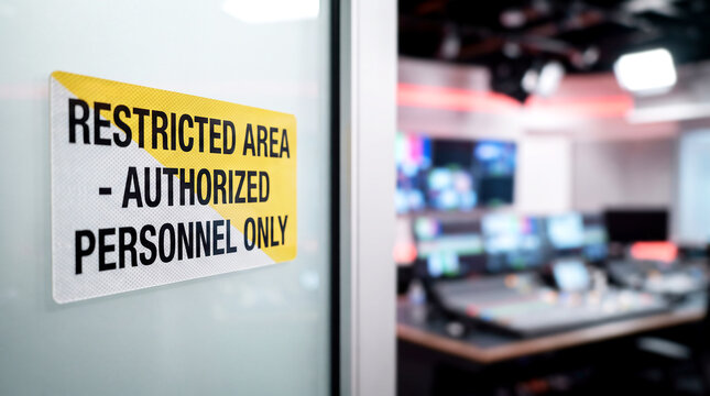 Warning sign on glass door leading to blurred television broadcast control room with monitors, studio security concept