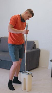 Young adult male wearing orange t shirt and blue shorts uses mop to clean floor inside. Man stands next to gray sofa holding onto handle of cleaning tool while focusing on his work