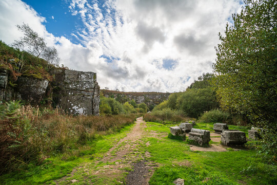 View of the historic Kit Hill Quarry with stone blocks scattered along a grassy path, surrounded by rocky cliffs and lush vegetation under a cloudy sky in Callington, England, United Kingdom.
