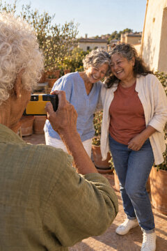 Senior friends laugh as one woman photographs them on a terrace