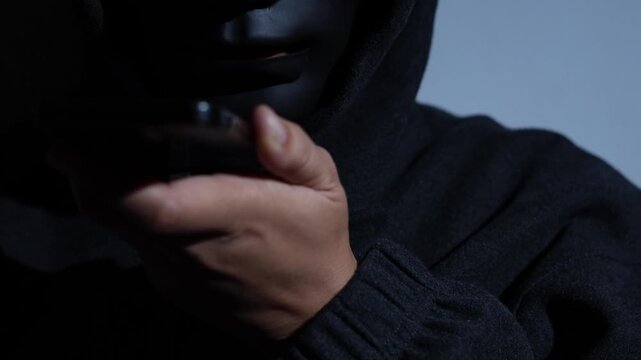 Phone scammers protection. Unknown businessman wearing mask with covered face using mobile makes an anonymous call intimidating and threatening the interlocutor on dark background. hacker call center.