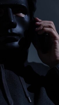 Phone scammers protection. Unknown businessman wearing mask with covered face using mobile makes an anonymous call intimidating and threatening the interlocutor on dark background. hacker call center.