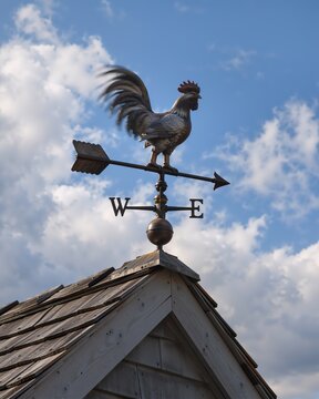 Weather vane with a rooster silhouette spinning on a wooden roof against a bright blue sky with scattered clouds, indicating wind direction and adding a rustic, charming element to the building's