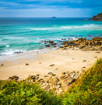 Lone surfer carrying a surfboard along a golden sand beach beside turquoise waves at Sennen Cove, Cornwall