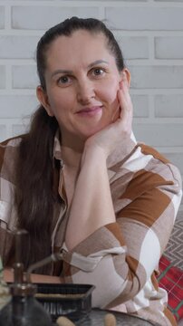 Woman rests her head on hand wearing brown checkered shirt against white brick wall background. Soft lighting illuminates red plaid fabric black tray with pump top visible in foreground