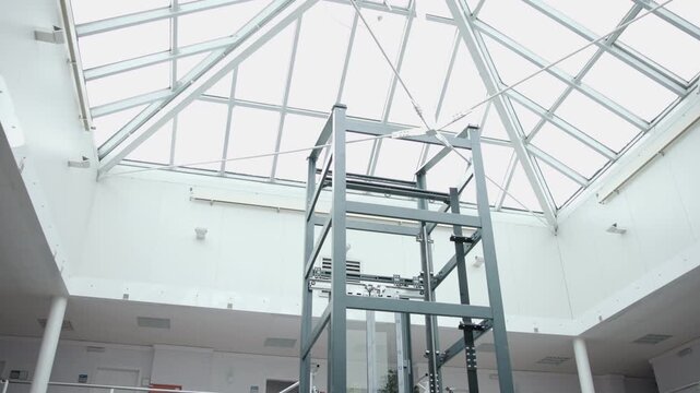 Industrial lift structure under pyramidal skylight in modern atrium with diffuse daylight, vertical cables, and diagonal trusses for mechanical inspection