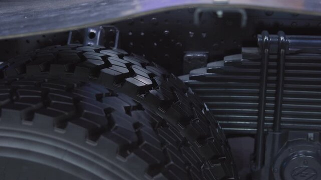 Low-angle close-up of dual heavy-duty truck tires and leaf spring suspension inside wheel well, showing chunky tread blocks, rubber texture, and blue directional lighting highlights