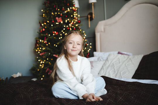 Kid girl wearing white knitted sweater sitting in bed over Christmas tree with glowing lights close up. Looking at camera. Winter holidays.