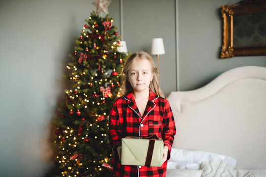 Kid girl 7-8 year old wearing red checkered pajamas holding Christmas present box over decorated eve in bedroom. Childhood. Winter holidays.