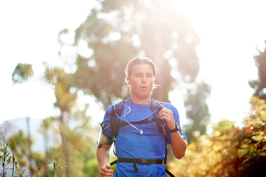 Fitness, man and running with earphones in forest for marathon playlist, breathing exercise and cardio music. Runner, sunlight and backpack on nature trail for workout performance or training podcast