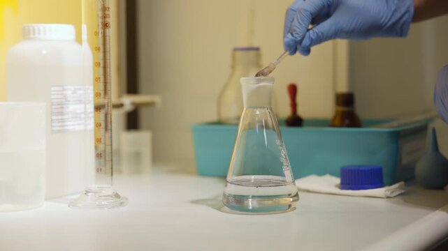 Gloved hand lowering small spoon to add reagent into Erlenmeyer flask with clear liquid on laboratory bench under warm overhead light, tabletop closeup, three quarter angle