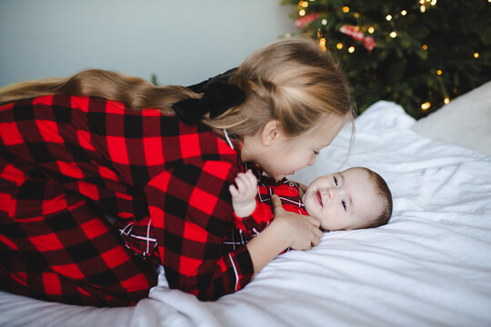 Two children playing in bed wearing red checkered pajamas having fun over Christmas tree at background in room. Winter holiday season.