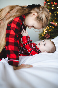 Child girl playing with baby in bed wearing red pajamas over Christmas tree at background. Winter holiday season. Childhood.