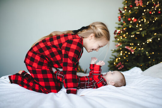 Child girl playing with baby in bed wearing red pajamas over Christmas tree at background. Winter holiday season. Childhood.