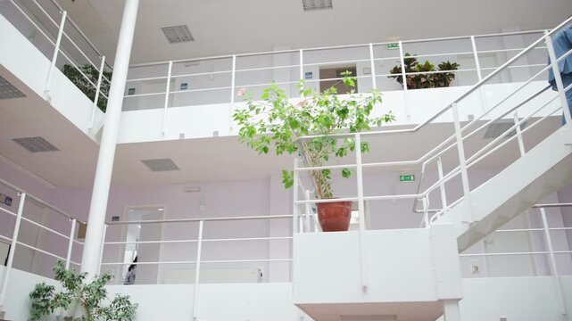 Businessman ascending white metal staircase in bright modern office atrium with potted plants, open mezzanine, and pastel interior walls