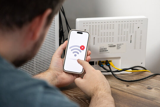 A person checking a smartphone with a no internet signal icon displayed on the screen near a white home router, depicting technical difficulties and connectivity issues at a residence