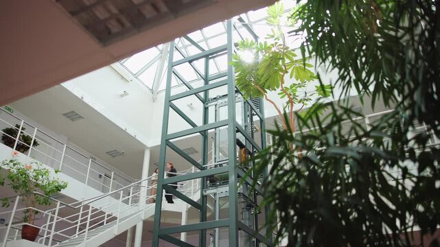 Bright office atrium interior with people walking by steel elevator shaft under glass skylight and diagonal stairs