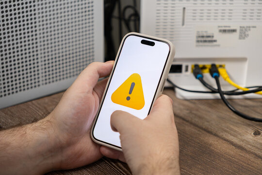 A person holds a smartphone displaying an alert icon near a home router, illustrating connectivity issues, network troubleshooting, and technical difficulties with internet signal