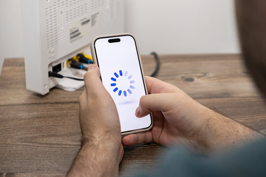 Person holding a smartphone with a circular blue loading symbol on the white screen near a home router, illustrating slow internet connectivity and network signal buffering issues daily