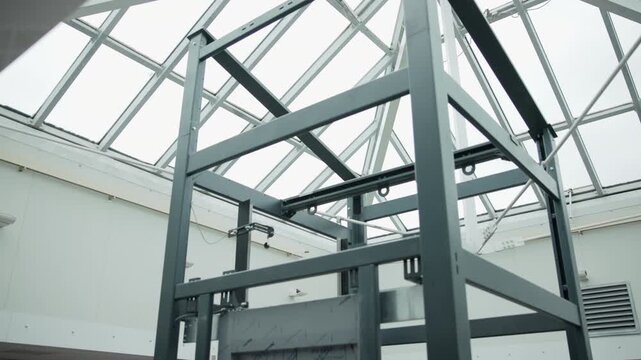 Upward low-angle view of dark steel framework with vertical posts, crossbeams, diagonal braces, and welded joints beneath glazed skylight in industrial atrium