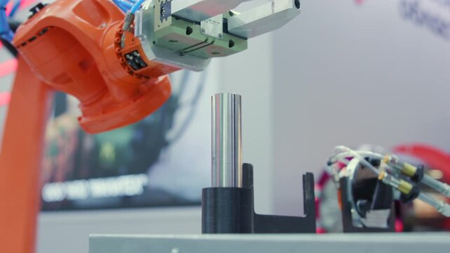Orange industrial robot arm gripper aligning above upright cylindrical steel post mounted in black tabletop fixture at trade show demonstration precision automation setup in showroom lighting
