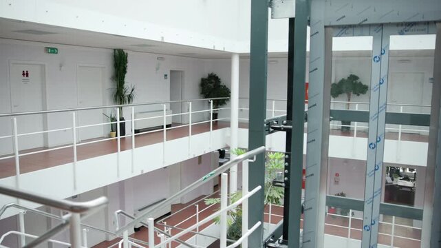 Bright modern atrium with glass elevator shaft, white railings, indoor plants, and open walkways in clean corporate building interior with natural daylight and architectural symmetry