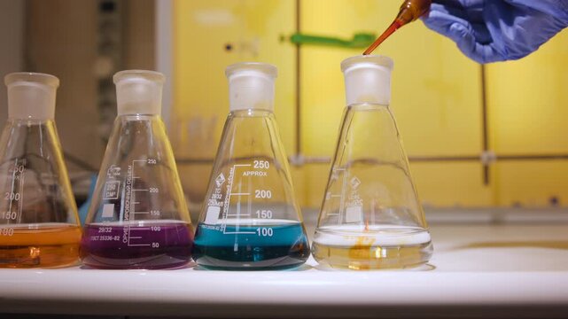 Gloved hand adding amber reagent drop from dropper over four colored Erlenmeyer flasks inside warm laboratory fume hood with visible droplet tip