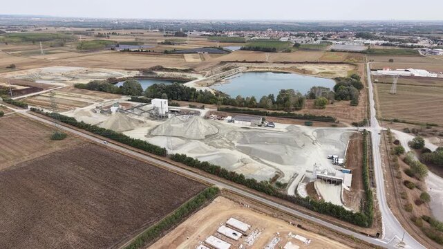 Aerial view of gravel quarry with piles of sand and industrial machinery near a lake in Chivasso, Piedmont, Italy.