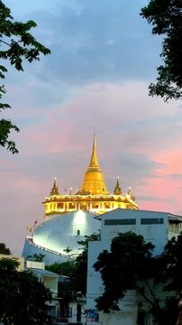 Golden Mountain Pagoda at Wat Saket standing beneath a vibrant pastel sunset sky with pink and blue clouds in Bangkok, Thailand, framed by silhouettes of lush green tree leaves in the evening.