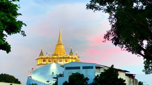 Golden Mountain Pagoda at Wat Saket standing beneath a vibrant pastel sunset sky with pink and blue clouds in Bangkok, Thailand, framed by silhouettes of lush green tree leaves in the evening.