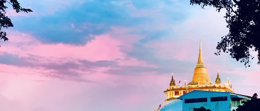 Golden Mountain Pagoda at Wat Saket standing beneath a vibrant pastel sunset sky with pink and blue clouds in Bangkok, Thailand, framed by silhouettes of lush green tree leaves in the evening.