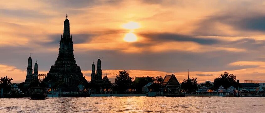 Silhouette of Wat Arun pagoda against a dramatic orange sunset sky over the Chao Phraya River in Bangkok, Thailand, creating a serene, golden twilight scene along the historic waterfront.