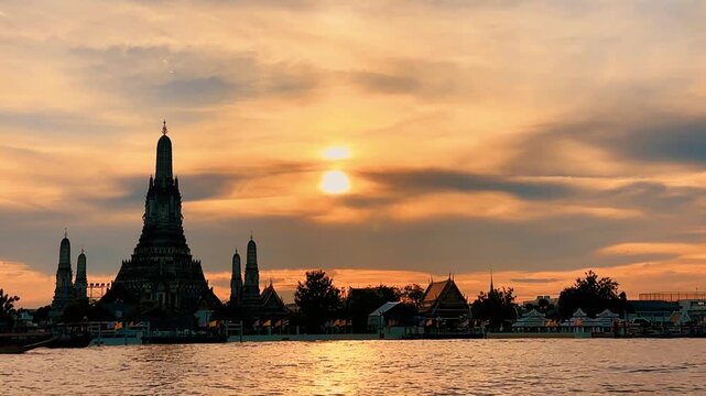 Silhouette of Wat Arun pagoda against a dramatic orange sunset sky over the Chao Phraya River in Bangkok, Thailand, creating a serene, golden twilight scene along the historic waterfront.
