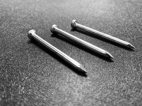 A precise, high-angle macro photograph of three common steel wire nails resting in a perfect parallel vertical alignment on a dark, rough-textured surface. Studio direct lighting. Suitable for industr