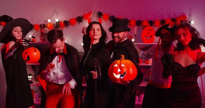 Happy friends celebrating Halloween party, spooky decorations, pumpkins, red lights, diverse group of funny cute people wearing costumes, dancing smiling to enjoy festive atmosphere, celebration