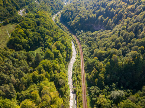 Railway follows river through forested gorge in Romania, with parallel tracks curving between wooded slopes and pale rocky stream. Aerial landscape highlights remote mountain rail access.