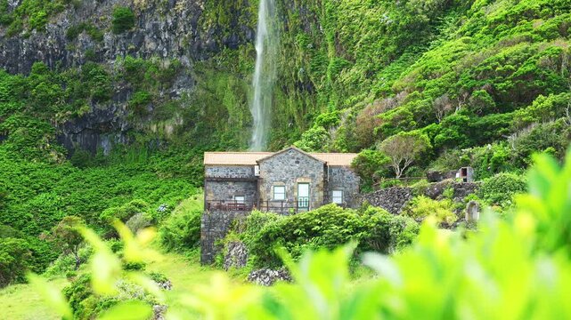 Wide scenic view of Cascata do Poco do Bacalhau waterfall flowing down a volcanic cliff behind a traditional stone house, framed by bright green foliage in Faja Grande, Flores Island. Rack focus