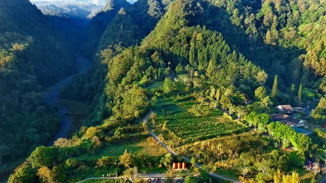 Aerial view of Kaliurang with lush green forests, a winding river, and small buildings nestled in the valley in Pakem, Special Region of Yogyakarta, Indonesia.