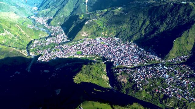 Aerial drone footage surveys green cultivated fields and crop rows across hilly rural terrain in Tungurahua, Ecuador. A winding road curves between the fields and scattered buildings, with strong sunl