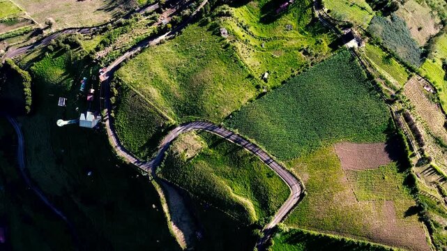 Drone footage shows a steep green mountain ridge in Tungurahua, Ecuador, with cultivated fields, winding roads, scattered buildings and communication towers along the slopes. Low clouds cling to the s