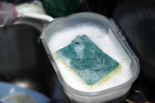 A green washing sponge resting in soapy white foam inside a clear plastic container near a kitchen sink