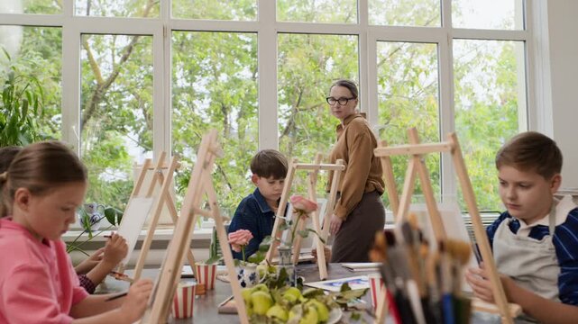 Caucasian female teacher supervising Caucasian children painting on easels during art workshop. Perfect for promoting creative educational courses, school classes, and tutoring programs