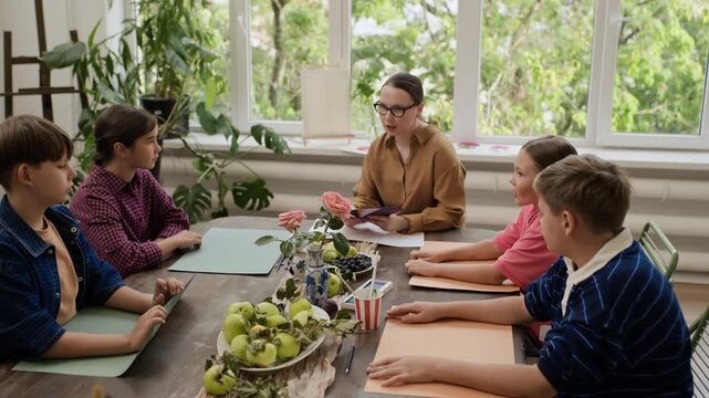 Caucasian female teacher showing color palette paper samples to group of children during art master class, zooming out. Visual fits creative workshop and art school advertising