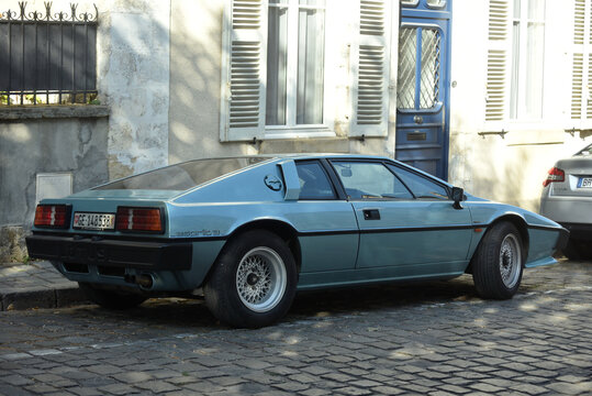 Bourges - France - 4 October 2026 - Rear three-quarter view of a light blue Lotus Esprit S3 sports car parked on a cobblestone street beside a weathered building with white shutters and a blue door.
