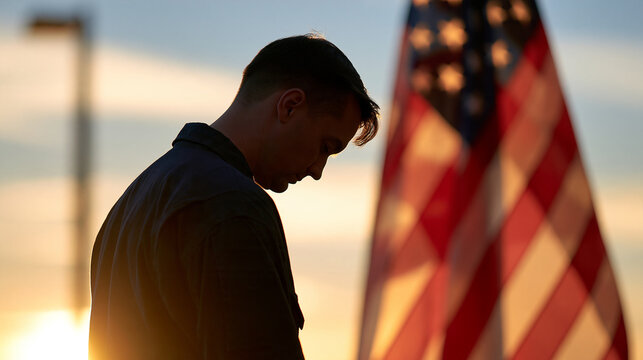 Faceless silhouette of a person standing with head bowed before an American flag at dawn, quiet remembrance, with copy space