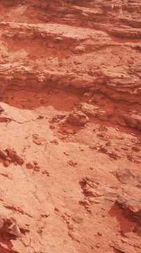 The scene shows a red rock area with various formations and textures. It appears dry with little to no vegetation. The sunlight highlights the intricate details in the rock surface.