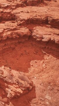The scene shows a dry area with red rocky ground and layered stone formations under bright sunlight. There are no plants or water visible in this barren landscape.