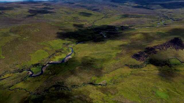 Drone footage presents rolling highland terrain in the Andes of Ecuador, where a narrow winding road crosses muted green and brown hills. Low clouds hang over distant mountain ridges beneath patches o