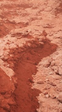 The scene shows a rocky red landscape with cracks and uneven ground. The sunlight casts long shadows across the surface. This place has a rugged and arid appearance typical of desert areas.